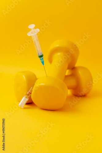syringe stuck in a dumbbell, a medical ampoule lies next to it, a yellow background, a vertical snapshot. the concept of doping in sports, steroids, testosterone and other drugs banned in sports.