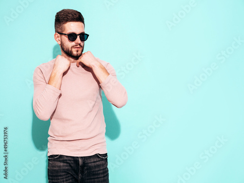 Portrait of handsome confident model. Sexy stylish man dressed in pink sweater and jeans. Fashion hipster male posing near blue wall in studio. In sunglasses. Isolated