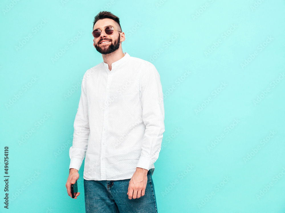 Portrait of handsome smiling model. Sexy stylish man dressed in white shirt and jeans. Fashion hipster male posing near blue wall in studio. Isolated. In sunglasses Portrait of handsome smiling model. Sexy stylish man dressed in white shirt and jeans. Fashion hipster male posing near blue wall in studio. Isolated. In sunglasses