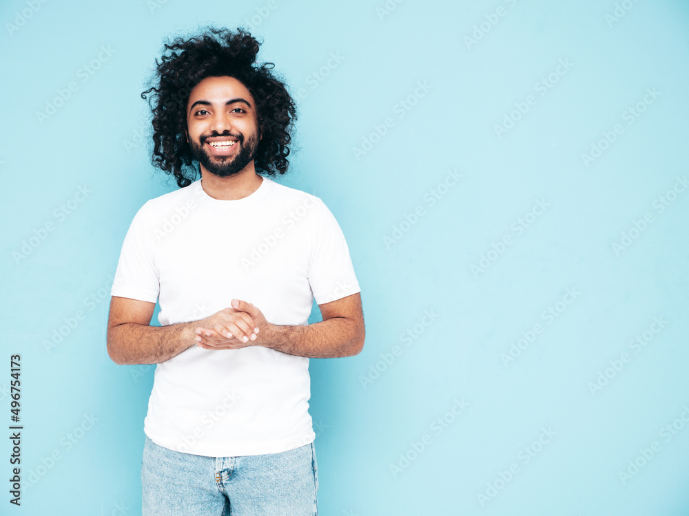 Handsome smiling hipster model.Sexy unshaven Arabian man dressed in white summer t-shirt and jeans clothes. Fashion male with long curly hairstyle posing near blue wall in studio. Isolated Handsome smiling hipster model.Sexy unshaven Arabian man dressed in white summer t-shirt and jeans clothes. Fashion male with long curly hairstyle posing near blue wall in studio. Isolated