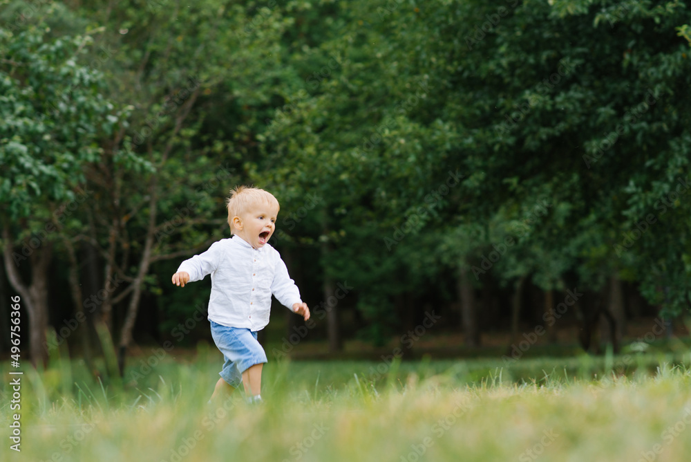 A little boy joyfully runs through the green grass in the summer in the park along a narrow path and smiles happily