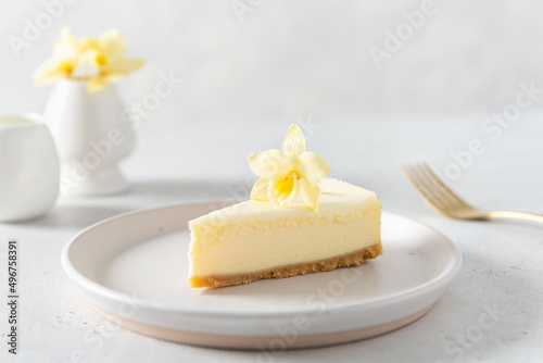 Classic New York cheesecake with fresh vanilla flower on white concrete background, side view. A piece of Vanilla cheesecake on a white plate. Confectionery menu, recipe. Close up.