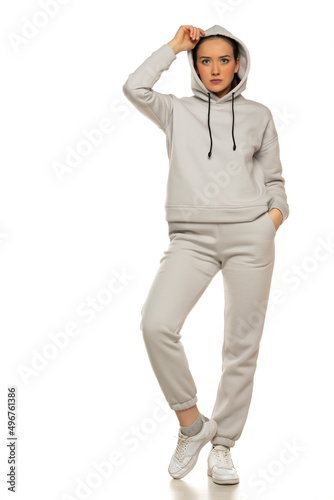 Front view of a young woman in a gray tracksuit and hood posing to a white background in the studio