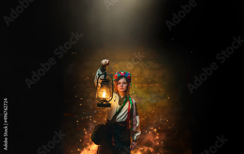 The woman in traditional, national clothes, a wreath with flowers on her head. In hands holds an old gas lamp. Illuminates a narrow passage in the dark basement, bomb shelter. Ukraine. Copy space.