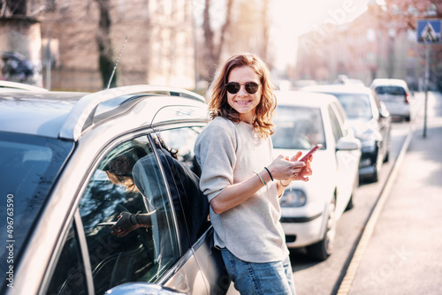 Happy cheerful young woman in sunglasses looking at the smartphone screen while standing by her gray car on the street