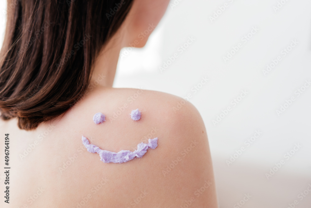 Smiley face made of purple cream scrub on shoulder of unrecognizable woman indoors. Skincare concept