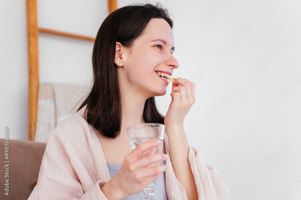 Smiling young woman holding pill omega capsule and glass of water looking away indoors. Girl taking medicine at home Smiling young woman holding pill omega capsule and glass of water looking away indoors. Girl taking medicine at home