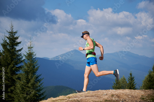 Light-skinned male runner of trail with sports bag on his waist and cap running from stone top of mountain hill. Healthy man jogging downhill among the summer nice mountain scenery.