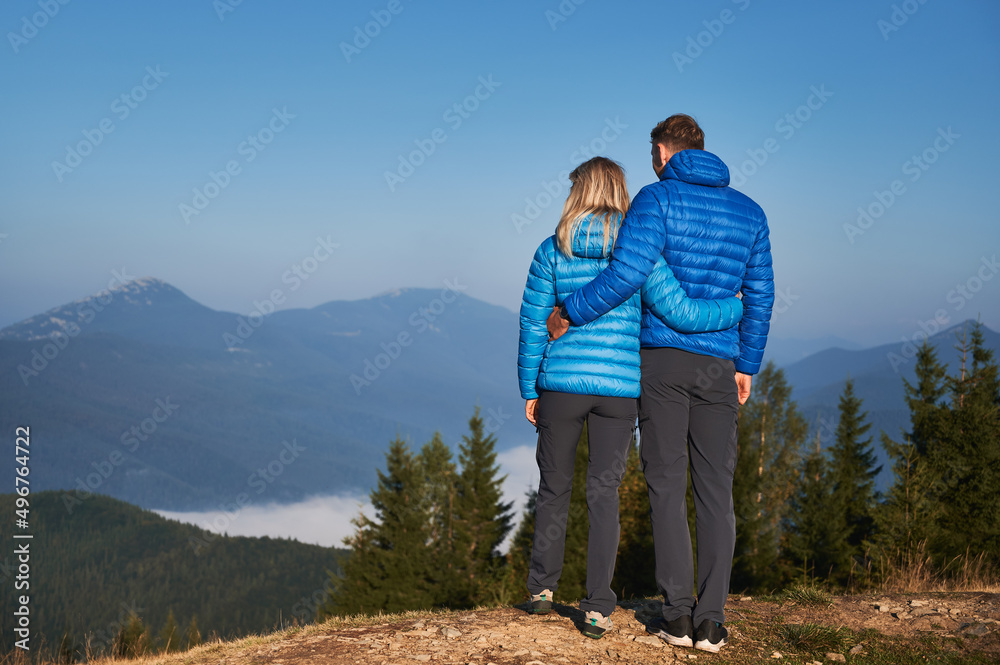 Back full length view of couple hikers holding each other around waist, contemplating on fabulous mountain landscape in front of them at sunshine autumn vacation.