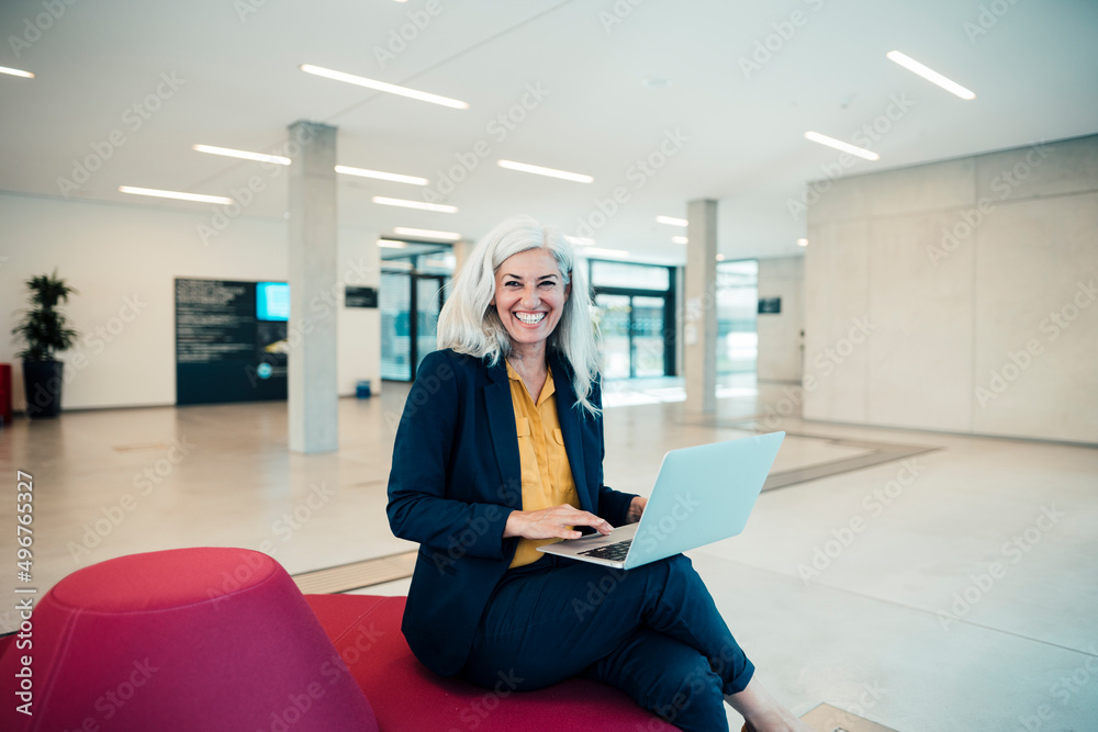 Cheerful businesswoman with laptop sitting on seat in office Cheerful businesswoman with laptop sitting on seat in office