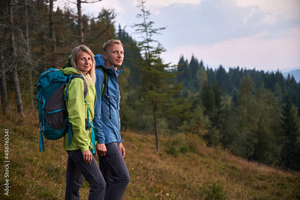 Portrait of man and woman with hiking equipment holding hands together, preparing start hike in the mountains. Green grass and coniferous forest on the background. Concept of hiking, relationships.