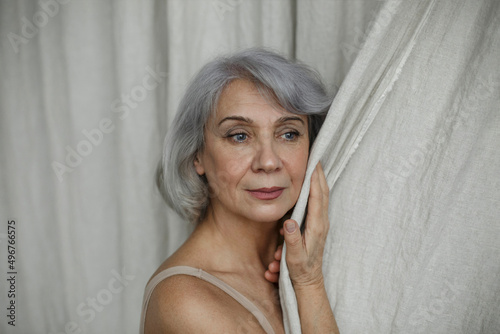 Thoughtful senior woman with gray hair touching curtain at home photo