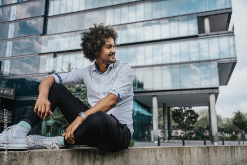 Smiling young man sitting on ledge in front of modern building