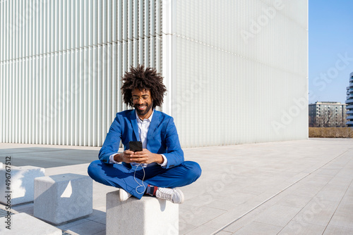 Smiling businessman listening music and using smart phone sitting on concrete block photo