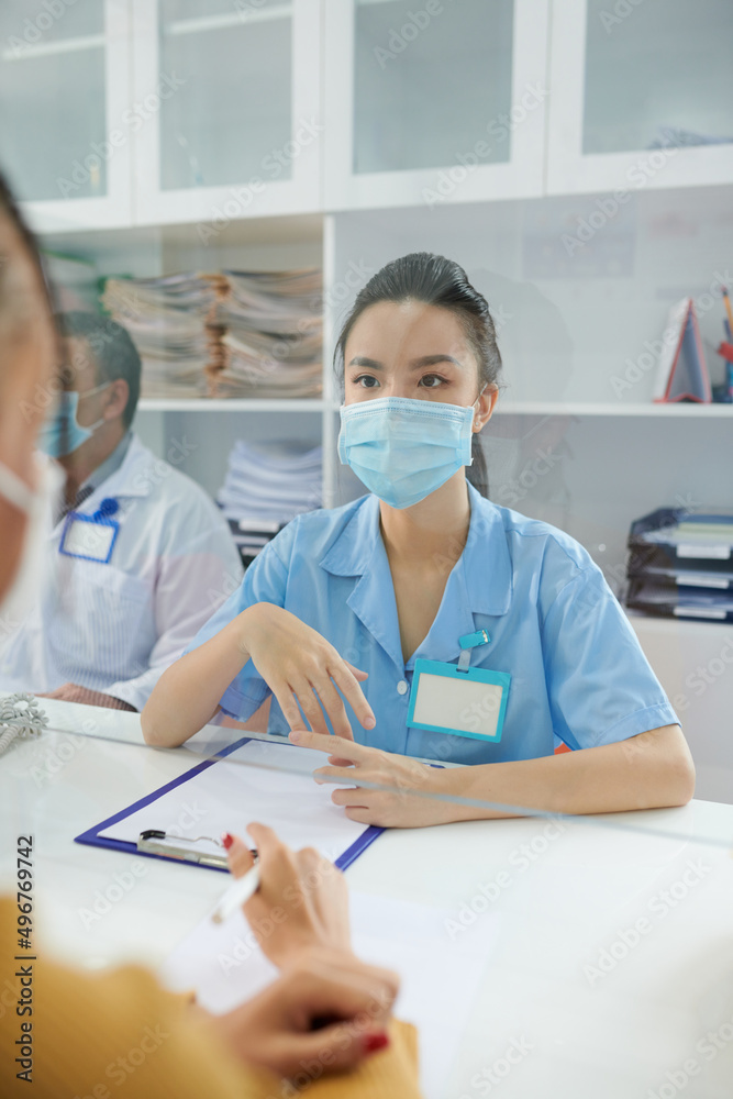 Young medical nurse in protective mask working at hospital reception, talking to patients and filling documents Young medical nurse in protective mask working at hospital reception, talking to patients and filling documents