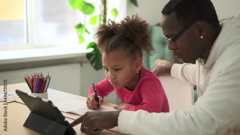 Little girl studying in online school and dad helping her, sitting at table at home room spbd. Closeup view of american african child looks at screen of tablet and writes in notebook, man helps her Little girl studying in online school and dad helping her, sitting at table at home room spbd. Closeup view of american african child looks at screen of tablet and writes in notebook, man helps her