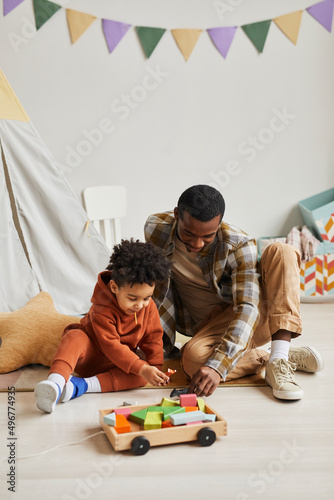 Vertical portrait of black father and son playing with toys together in cute kids room interior