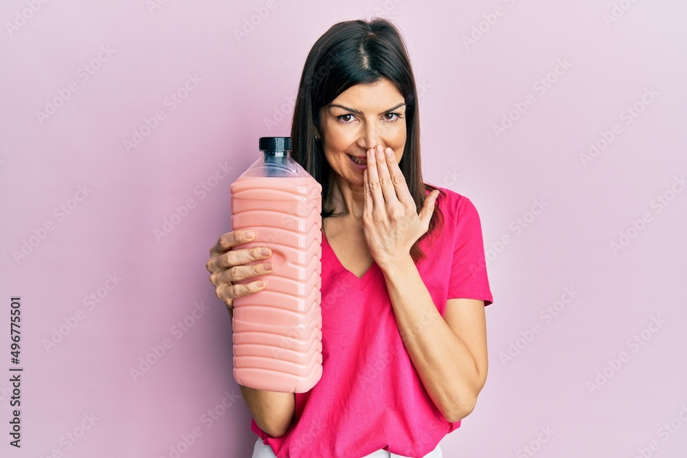 Young hispanic woman holding laundry softener bottle covering mouth with hand, shocked and afraid for mistake. surprised expression