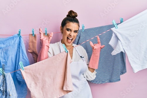 Beautiful brunette young woman washing clothes at clothesline shouting with crazy expression doing rock symbol with hands up. music star. heavy concept.