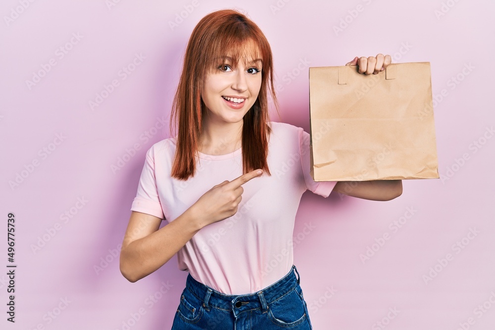 Redhead young woman holding take away paper bag smiling happy pointing with hand and finger Redhead young woman holding take away paper bag smiling happy pointing with hand and finger