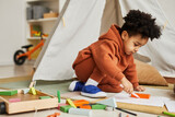 Full length portrait of cute toddler boy drawing on floor in kids room while dressed in trendy neutral outfit, copy space