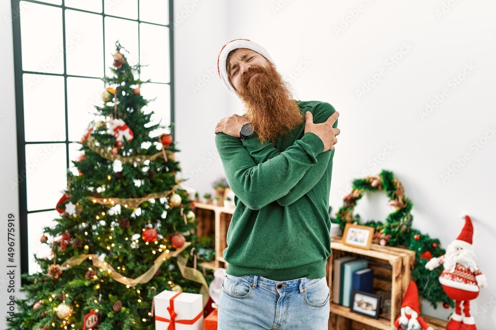 Redhead man with long beard wearing christmas hat by christmas tree hugging oneself happy and positive, smiling confident. self love and self care Redhead man with long beard wearing christmas hat by christmas tree hugging oneself happy and positive, smiling confident. self love and self care