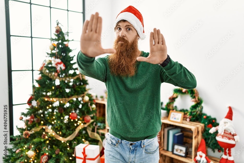 Redhead man with long beard wearing christmas hat by christmas tree doing frame using hands palms and fingers, camera perspective Redhead man with long beard wearing christmas hat by christmas tree doing frame using hands palms and fingers, camera perspective