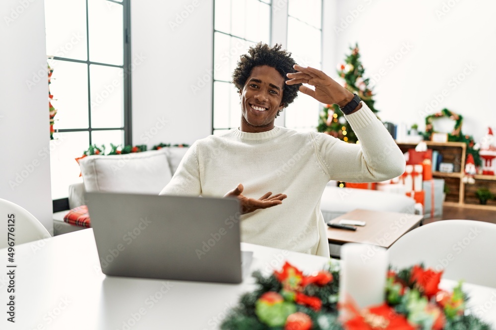 Young african american man using laptop sitting on the table by christmas tree gesturing with hands showing big and large size sign, measure symbol. smiling looking at the camera. measuring concept.