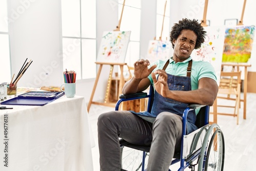 Young african american artist man sitting on wheelchair at art studio disgusted expression, displeased and fearful doing disgust face because aversion reaction. with hands raised