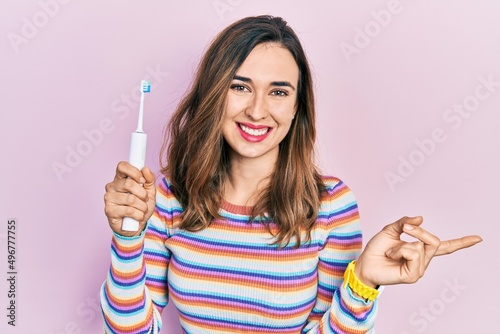 Young hispanic girl holding electric toothbrush smiling happy pointing with hand and finger to the side