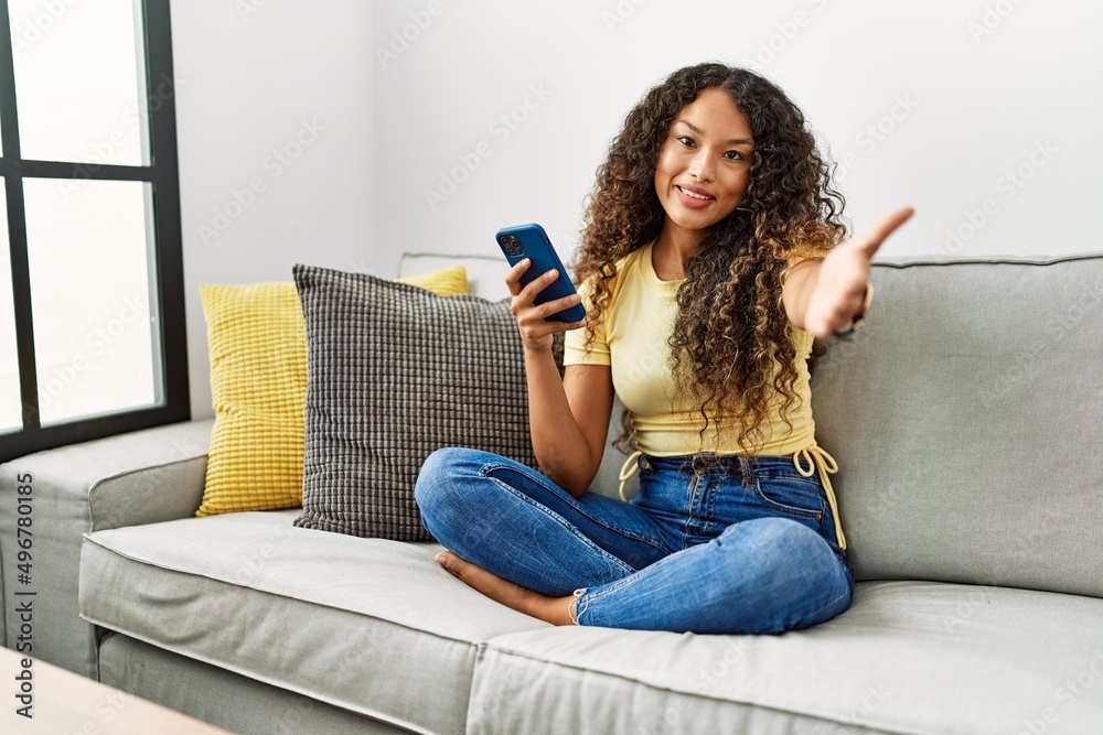 Beautiful hispanic woman sitting on the sofa at home using smartphone smiling friendly offering handshake as greeting and welcoming. successful business. Beautiful hispanic woman sitting on the sofa at home using smartphone smiling friendly offering handshake as greeting and welcoming. successful business.