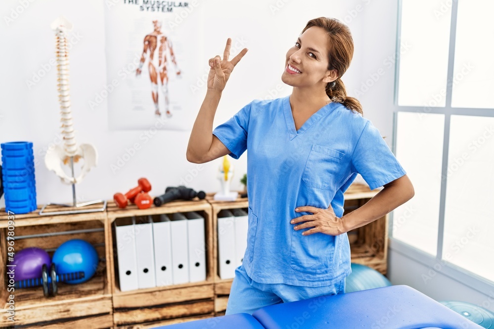 Middle age hispanic physiotherapist woman working at pain recovery clinic smiling looking to the camera showing fingers doing victory sign. number two. Middle age hispanic physiotherapist woman working at pain recovery clinic smiling looking to the camera showing fingers doing victory sign. number two.