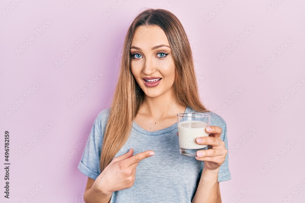 Young blonde girl holding glass of milk smiling happy pointing with hand and finger Young blonde girl holding glass of milk smiling happy pointing with hand and finger