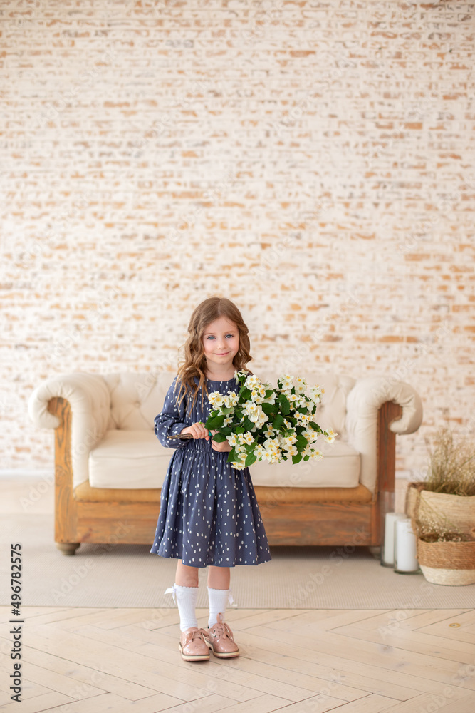 Smiling little curly hair girl with a bouquet of jasmine flowers stands near sofa in a cozy living room. Scandinavian interior. Celebration. Mothers Day. Child with a bouquet of flowers for mom. Smiling little curly hair girl with a bouquet of jasmine flowers stands near sofa in a cozy living room. Scandinavian interior. Celebration. Mothers Day. Child with a bouquet of flowers for mom.
