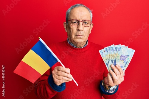Handsome senior man with grey hair holding romania flag and leu banknotes relaxed with serious expression on face. simple and natural looking at the camera.
