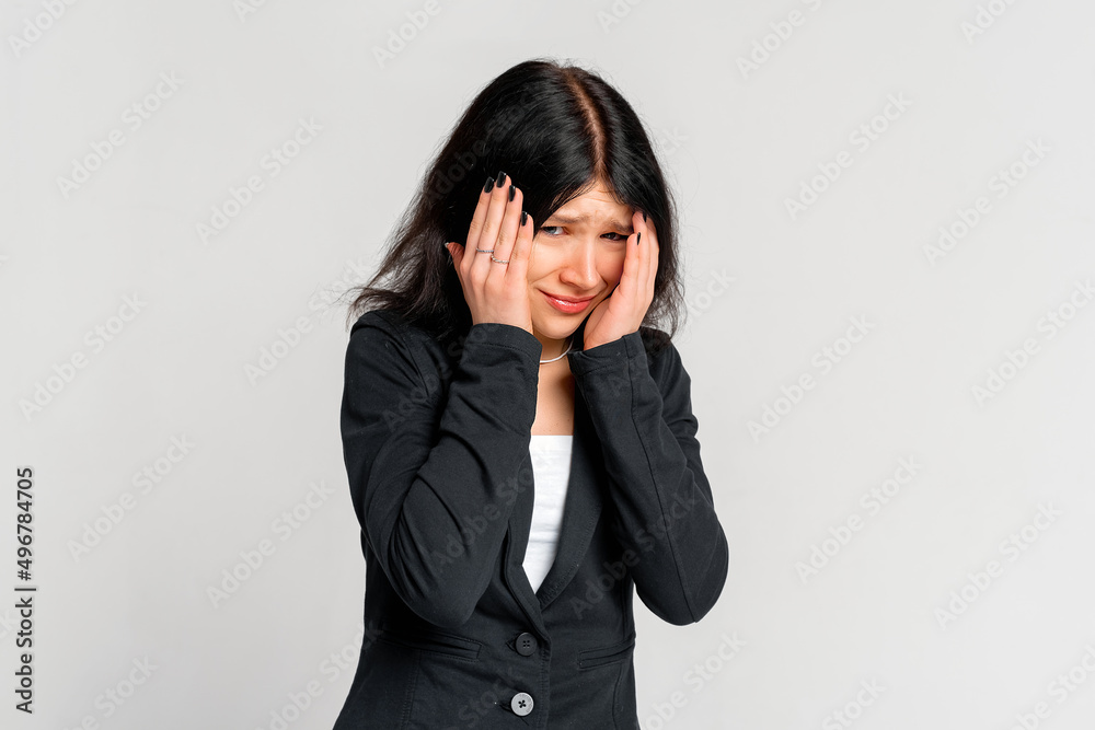 Portrait of startled young brunette woman grabbing head with both hands in despair , staring shocked and confused at camera, standing in black blazer over grey background Portrait of startled young brunette woman grabbing head with both hands in despair , staring shocked and confused at camera, standing in black blazer over grey background