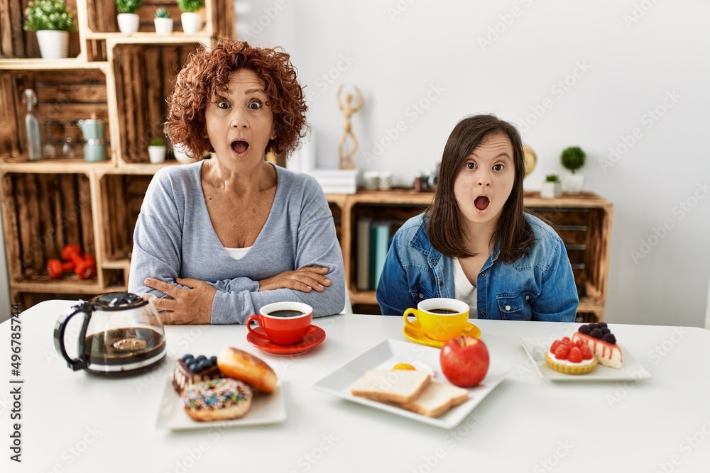 Family of mother and down syndrome daughter sitting at home eating breakfast afraid and shocked with surprise expression, fear and excited face.