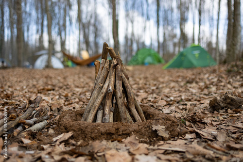Unburned campfire with tree branches in forest. Autumn leaves on ground. Camping tents and hammock at blur background.