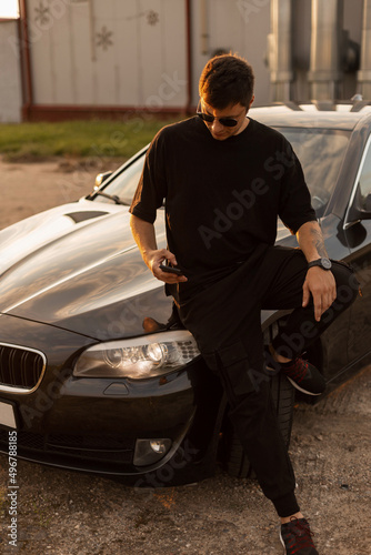 Fashionable handsome businessman hipster guy with cool sunglasses in black T-shirt and jeans sitting near car and watching a smartphone at sunset