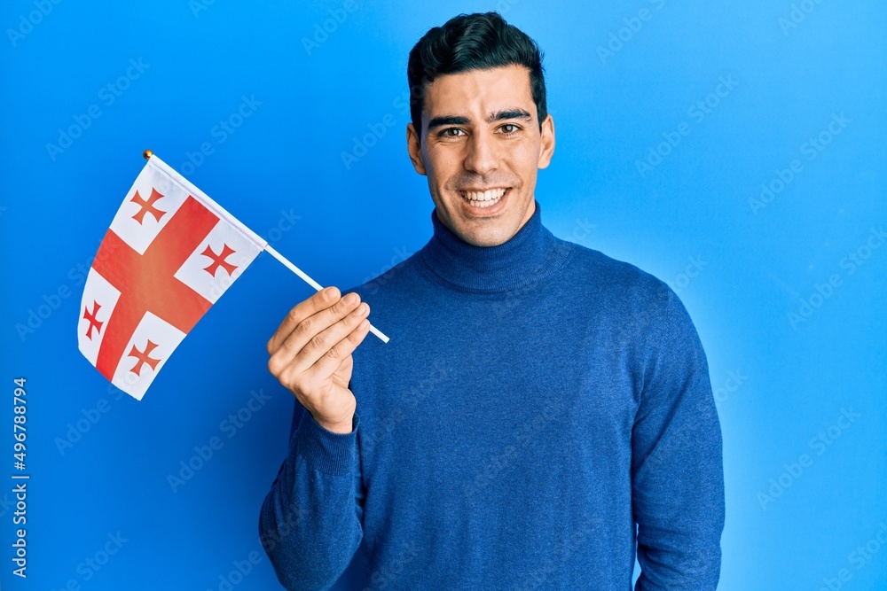 Handsome hispanic man holding georgia flag looking positive and happy standing and smiling with a confident smile showing teeth