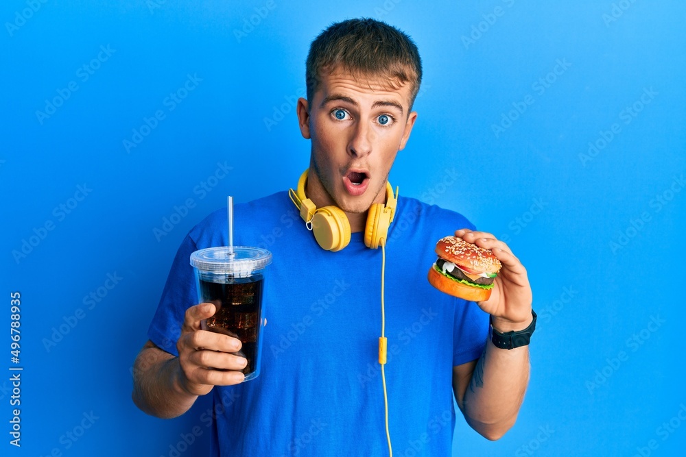 Young caucasian man eating a tasty classic burger and soda afraid and shocked with surprise and amazed expression, fear and excited face. Young caucasian man eating a tasty classic burger and soda afraid and shocked with surprise and amazed expression, fear and excited face.