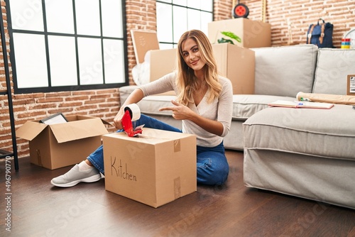 Young blonde woman sitting on the floor closing boxes home smiling happy pointing with hand and finger