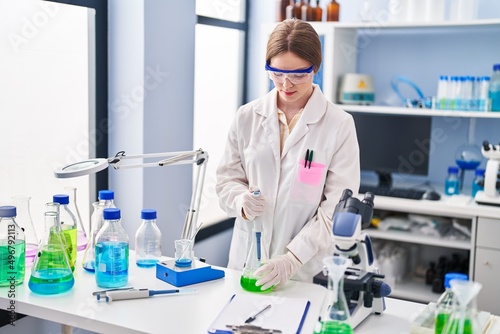 Young blonde woman wearing scientist uniform measuring liquid on test tube at laboratory