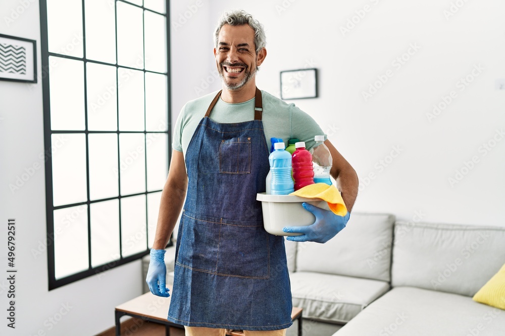 Middle age hispanic man wearing cleaner apron holding cleaning products looking positive and happy standing and smiling with a confident smile showing teeth