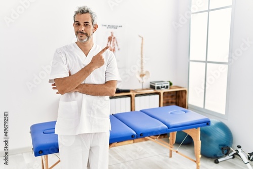 Middle age hispanic therapist man working at pain recovery clinic pointing with hand finger to the side showing advertisement  serious and calm face