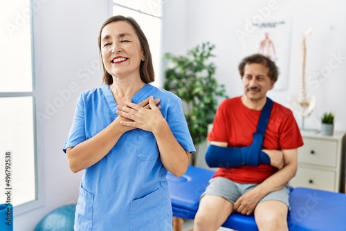 Middle age doctor woman with patient with arm injury at rehabilitation clinic smiling with hands on chest  eyes closed with grateful gesture on face. health concept.