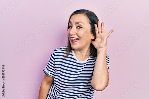 Middle age hispanic woman wearing casual clothes smiling with hand over ear listening and hearing to rumor or gossip. deafness concept.