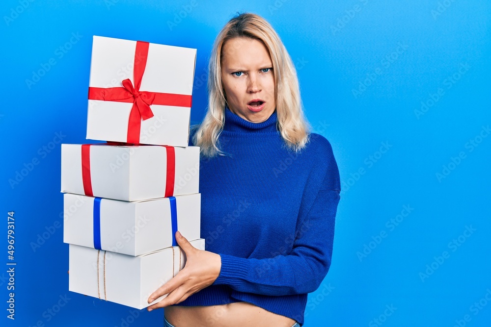 Beautiful caucasian blonde woman holding gifts in shock face, looking skeptical and sarcastic, surprised with open mouth Beautiful caucasian blonde woman holding gifts in shock face, looking skeptical and sarcastic, surprised with open mouth