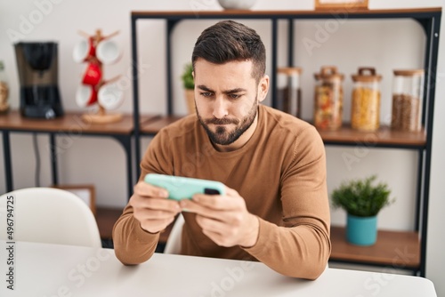Young hispanic man using smartphone sitting on table at home