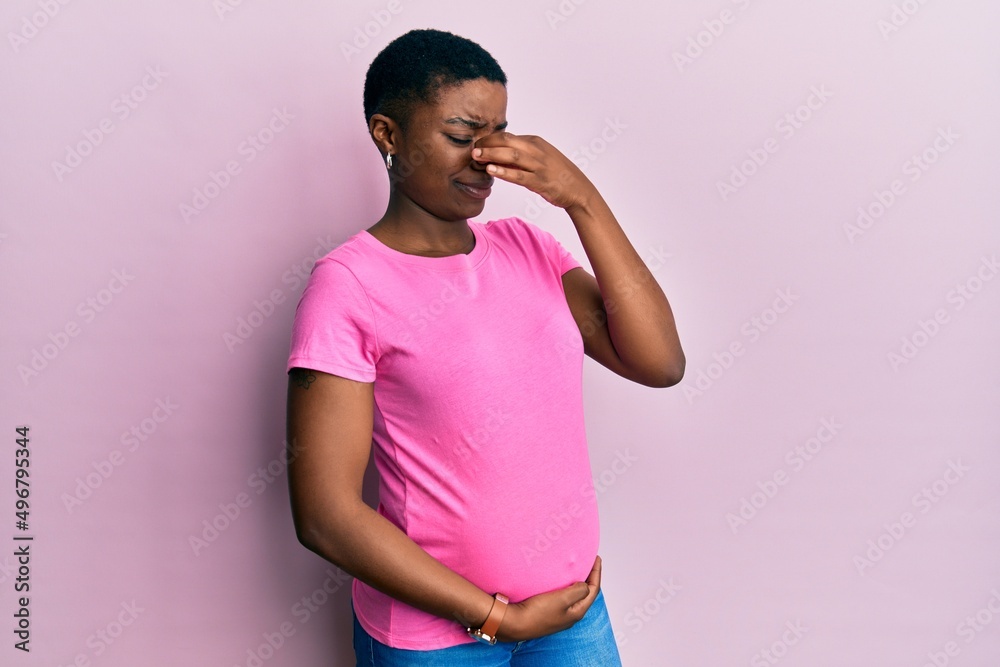 Young african american woman expecting a baby, touching pregnant belly smelling something stinky and disgusting, intolerable smell, holding breath with fingers on nose. bad smell